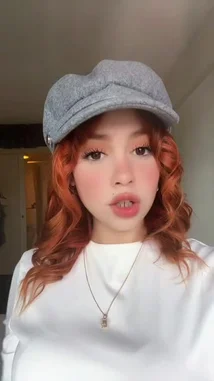 A young woman with vibrant orange hair and a stylish gray cap showcases her look in a casual selfie-style video. She wears a white top and a delicate necklace, making direct eye contact with the camera. The setting appears to be a bedroom, creating an intimate and personal feel. by @merlinblok3 with Original Sound by merlinblok3