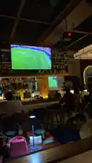 In a vibrant sports bar buzzing with excitement, Everything Naddy is captured in a moment of pure joy. She cheers loudly while wearing a Ghana national team jersey and holding a festive green cocktail, her face lit up with a smile as she celebrates what sounds like a goal during a live soccer match on the big screen. by @everythingnaddy with Original Sound by everythingnaddy