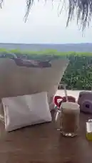 Experience a serene day at a tropical beachside retreat. The scene opens with a stylish table setting under a thatched roof, featuring a refreshing drink, a pink instant camera, and a woven tote bag, all overlooking a lush green hedge and the calm ocean. As the camera pans, it reveals a tranquil patio area with wooden furniture and palm trees swaying gently in the breeze, capturing the essence of island relaxation. by @codedsoso with Original Sound by codedsoso