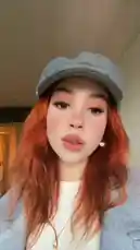 A young woman with vibrant orange-red hair showcases her stylish outfit, featuring a grey newsboy cap, a white top, and a light blue coat. She speaks directly to the camera in a casual indoor setting, presenting a simple yet chic fashion look. by @merlinblok3 with Original Sound by merlinblok3