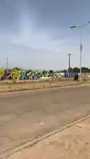 This eye-level journey captures a vibrant stretch of roadside wall art under a clear sky. A colorful auto-rickshaw passes by, adding life to the scene as the camera moves along the pavement. The mural-covered wall features bold patterns and text, suggesting a rich local artistic community. This authentic glimpse into everyday street culture is perfect for those interested in travel, photography, and urban exploration. by @fdlsage with fdlsage by Original Sound