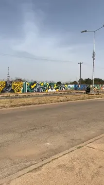 This eye-level journey captures a vibrant stretch of roadside wall art under a clear sky. A colorful auto-rickshaw passes by, adding life to the scene as the camera moves along the pavement. The mural-covered wall features bold patterns and text, suggesting a rich local artistic community. This authentic glimpse into everyday street culture is perfect for those interested in travel, photography, and urban exploration. by @fdlsage with fdlsage by Original Sound