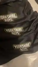 This close-up walkthrough reveals the details of new 'Yorkshire Boys' apparel from PACK'D. The camera focuses on the black fabric with its distinctive white and green logo, then moves to show the product's packaging, which features 'Let's Ride By All Means' text and various activity icons. The clear plastic bag displays 'MADE IN CHINA,' providing insight into the product's origin. by @pack.d with Original Sound by Contains: See You Again - Double M