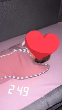 A baby peacefully sleeping in a pink swaddle blanket inside a crib. The scene is calm and serene, with soft lighting and a digital clock showing 2:49 in the background. A large red heart emoji is overlaid on the image, adding a touch of affection and warmth to the moment. by @framesofbrum with Original Sound by framesofbrum
