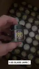 A close-up showcase of custom-designed product jars with vibrant 'SFC' branding. The video highlights the intricate purple and yellow graphic design on black lids, presented in bulk packaging. A hand demonstrates the product, emphasizing its unique aesthetic and potential for branding. by @pack.d with Left Hollywood by Northsidebenji