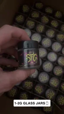 A close-up showcase of custom-designed product jars with vibrant 'SFC' branding. The video highlights the intricate purple and yellow graphic design on black lids, presented in bulk packaging. A hand demonstrates the product, emphasizing its unique aesthetic and potential for branding. by @pack.d with Left Hollywood by Northsidebenji