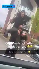 A man in a black hoodie and beanie playfully carries another man on his shoulders while holding a shovel. They walk down a street in what appears to be a lighthearted, comedic moment between friends. The scene has a candid, 'real life' feel, capturing a spontaneous interaction. by @uk-gos with Original Sound by uk-gos