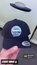 A hand holds and displays a black trucker hat featuring the 'Man With A Van' logo. The hat has a circular patch with a cartoon drawing of a man next to a white van, with the text 'MAN WITH A VAN' below it. The background shows a light-colored vehicle door handle. In the bottom right corner, a music player overlay shows the song 'Wanksta' by 50 Cent. by @pack.d with Original Sound by Contains: Wanksta (From "8 Mile" Soundtrack) - 50 Cent