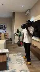 A man in a casual white t-shirt, black pants, and yellow shoes demonstrates a creative home workout using a bowling ball as a weight. He holds the ball in one hand while juggling a red cup with the other, showcasing strength and coordination. The setting is a modern living room with hardwood floors, a large TV, and contemporary furniture, illustrating an innovative way to exercise without traditional gym equipment. by @fdlsage with fdlsage by Original Sound