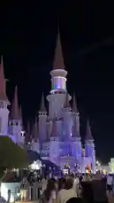 A vibrant night scene captures a large crowd gathered in front of a majestic, illuminated castle resembling Cinderella's Castle. The castle's spires are lit with dynamic purple and blue lights, creating a magical atmosphere. The soundscape features epic, cinematic music, enhancing the sense of wonder and spectacle. by @n.c7x with Original Sound by n.c7x