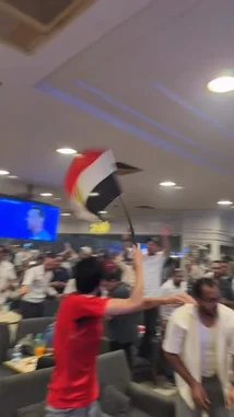 A group of Egyptian soccer fans erupt in celebration inside a restaurant after their team scores a goal during a World Cup match. The crowd cheers, waves the Egyptian flag, and films the moment on their phones as the players celebrate on the large screen. by @snap_alshle with Original Sound by snap_alshle