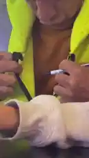 A man wearing a bright yellow high-visibility jacket is seen leaning over a dark counter, signing something with a black marker pen. A person in a white long-sleeved shirt is partially visible next to him. In the background, a red sign with the letters 'EN' can be seen. by @uk-gos with Original Sound by Contains: Marry me rasta - Maria mantereng