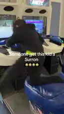 A young boy attempts to climb onto a racing arcade machine in an amusement center. He struggles to get on top, prompting on-screen text that reads 'Someone get this kid a Surron.' The scene captures a candid, humorous moment of playful mischief. by @uk-gos with Skrilla by Kodak Black