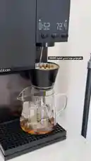 A close-up view shows a sleek black coffee machine brewing fresh iced coffee. Hot water is dispensed into a filter basket filled with ground coffee, which then drips through into a glass carafe containing ice cubes. The digital display on the machine counts down the brewing time, illustrating a simple and modern method for preparing a refreshing cold brew. by @alysf90 with Original Sound by alysf90