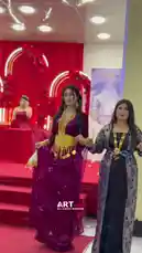 A group of women in elaborate traditional attire perform a synchronized dance on a red-carpeted stage. The setting features vibrant red backdrops with floral arches, suggesting a festive cultural or celebratory event. The performers wear ornate gold accessories and flowing garments, moving gracefully as part of a choreographed routine. by @artvideo.me with artvideo.me by Original Sound