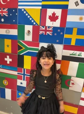 A young girl with black hair styled in pigtails stands smiling in front of a vibrant wall covered in various national flags. She is wearing a black dress with sheer polka-dot sleeves and a ruffled skirt. She poses confidently, then begins to dance playfully, spinning around and moving her arms joyfully. by @ufmradio with Original Sound by ufmradio