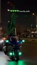 Witness the vibrant energy of a Saudi National Day celebration at night. The scene unfolds on a bustling city street illuminated by festive lights and crowds of people. A highlight of the event is a paraglider soaring through the dark sky, trailing a spectacular line of fire, creating a breathtaking spectacle for the onlookers below. by @wedad_23g with Original Sound by wedad_23g