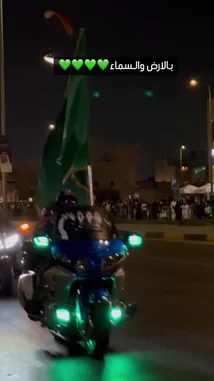 Witness the vibrant energy of a Saudi National Day celebration at night. The scene unfolds on a bustling city street illuminated by festive lights and crowds of people. A highlight of the event is a paraglider soaring through the dark sky, trailing a spectacular line of fire, creating a breathtaking spectacle for the onlookers below. by @wedad_23g with Original Sound by wedad_23g