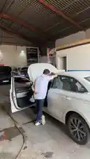 This walkthrough video provides a behind-the-scenes look at Wasta Hawkar Service, a professional auto repair garage. A technician is seen working on a white car with its trunk open, while other vehicles are serviced in the background. The setting features a corrugated metal roof, concrete floors, and prominent branding for 'Hawkar Service' and 'Liqui Moly,' highlighting the workshop's equipment and atmosphere. by @hawkar_service with Original Sound by hawkar_service