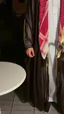 This video captures a moment of cultural display, featuring a person in traditional Saudi attire, including a red-and-white checkered keffiyeh and a brown bisht. A server presents a tray with a green espresso cup and saucer, bearing Arabic script. The centerpiece of the table is a large, ornate silver sword, which the individual then picks up to stir their coffee. They proceed to examine a small card with text and an image, possibly related to the sword or the event. The setting is dimly lit, suggesting an evening gathering. by @fox_cofee with Original Sound by fox_cofee