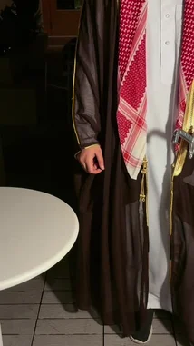 This video captures a moment of cultural display, featuring a person in traditional Saudi attire, including a red-and-white checkered keffiyeh and a brown bisht. A server presents a tray with a green espresso cup and saucer, bearing Arabic script. The centerpiece of the table is a large, ornate silver sword, which the individual then picks up to stir their coffee. They proceed to examine a small card with text and an image, possibly related to the sword or the event. The setting is dimly lit, suggesting an evening gathering. by @fox_cofee with Original Sound by fox_cofee