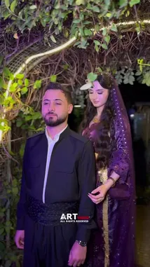 A beautifully lit outdoor scene captures a couple sharing an intimate embrace. The woman, adorned in a stunning purple sequined outfit and veil, wraps her arms around the man in a black traditional tunic. Set against a backdrop of lush greenery illuminated by golden string lights, their affectionate moment creates a romantic and cinematic atmosphere. by @artvideo.me with artvideo.me by Original Sound