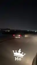 A silver car drives down a dark road at night, its brake lights glowing red. The camera follows from behind, capturing the vehicle's movement through an industrial area with large warehouses illuminated by overhead lights. A white crown icon with the word 'Me' is overlaid on the screen, suggesting the perspective of the driver. by @khllo17 with Original Sound by khllo17
