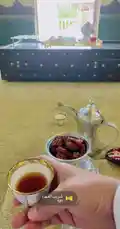 A close-up view of a hand holding a traditional Arabic coffee cup filled with dark, rich coffee. In the background, a silver dallah (coffee pot) and a bowl of fresh dates sit on a golden-colored tablecloth. The setting appears to be a cozy room with a window and decorative elements, suggesting a moment of hospitality or cultural tradition. by @s3432x with Original Sound by s3432x