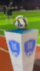 This video captures the vibrant atmosphere of the 'Al Jabal Al Gheeb' football tournament held at night. It begins with a close-up of the official tournament ball on its branded stand. The scene transitions to players from opposing teams, one in blue/yellow kits and the other in red/orange, lining up for the pre-match handshake. Officials, including referees in red shirts, are present on the field. A man speaks into a microphone near a 'Welcome' archway, while another person operates a professional camera on a tripod. Spectators watch from white couches and bleachers under bright stadium lights. The video concludes with live gameplay, showing players in action on the pitch, with various sponsor banners visible around the field. by @dalilnet with Original Sound by dalilnet