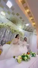 This elegant scene captures two brides in magnificent white wedding dresses posing together at a lavishly decorated reception. One bride wears a sparkling off-the-shoulder gown, while the other is in a voluminous ballgown with feathered sleeves. They stand amidst beautiful floral arrangements and ornate gold accents, creating a dreamy atmosphere perfect for a luxury wedding celebration. by @artvideo.me with artvideo.me by Original Sound
