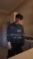 In a dimly lit kitchen, Badr Al-Qahtani performs a trending dance move. He is wearing a blue shirt with white Arabic text and black pants. A white cast is visible on his left hand. The background features white cabinets, a refrigerator, and a gas stove. The audio consists of a female voice singing in English, followed by a musical sting. by @cviioul with Original Sound by Contains: I Deserve to Bleed - Sushi Soucy