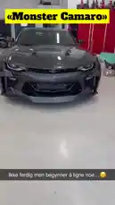 A sleek, dark grey Chevrolet Camaro with aggressive custom bodywork and black wheels is showcased inside a well-lit workshop. The camera pans around the vehicle, highlighting its modified front bumper, side skirts, rear spoiler, and overall wide-body aesthetic. In the background, red tool cabinets and other automotive equipment are visible. The car appears to be a work-in-progress, with some parts still taped off. The audio features a modern hip-hop track with a strong beat and male vocals. by @vikingcustom with GHOST OF NORWAY by RAVNLORE