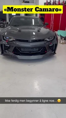 A sleek, dark grey Chevrolet Camaro with aggressive custom bodywork and black wheels is showcased inside a well-lit workshop. The camera pans around the vehicle, highlighting its modified front bumper, side skirts, rear spoiler, and overall wide-body aesthetic. In the background, red tool cabinets and other automotive equipment are visible. The car appears to be a work-in-progress, with some parts still taped off. The audio features a modern hip-hop track with a strong beat and male vocals. by @vikingcustom with GHOST OF NORWAY by RAVNLORE