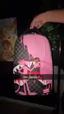 A hand presents a vibrant pink backpack featuring a prominent shark mouth design and a character resembling Sabrina Carpenter. The bag, which has a black and white checkered pattern on its side and a red 'Shark Head' tag, is shown from multiple angles against a dark background. by @absi_fashion with absi_fashion by Contains: Somebody - Latto