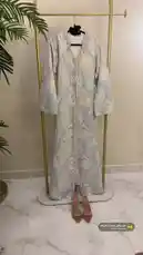A stunning light blue and cream marbled kaftan dress with intricate gold embroidery and pink beadwork is displayed on a modern gold clothing rack. The camera pans slowly down the full-length garment, highlighting its V-neckline, bell sleeves, and detailed front embellishments. A pair of matching brown strappy heels rests neatly on the floor below, completing the elegant ensemble. This presentation showcases the craftsmanship of traditional attire, perfect for formal events. by @needle_t with موسيقى جميلة ذكريات by MA Production