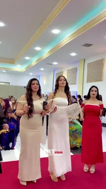 A group of elegantly dressed women walk confidently down a red carpet at what appears to be a formal event or celebration. They wear beautiful gowns in various colors including white, red, blue, and green, showcasing their fashion and poise. The setting is an indoor venue with guests seated at tables watching the procession. by @artvideo.me with artvideo.me by Original Sound