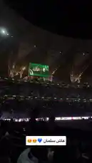 This immersive point-of-view footage captures the vibrant energy inside a massive, modern stadium during a major event in Saudi Arabia. The camera pans across thousands of spectators holding up their phones, creating a breathtaking sea of lights under the dark sky. A large digital screen displays Arabic text and logos, while dynamic purple and red stage lighting illuminates the field below. The roar of the crowd is palpable, providing an authentic sense of being part of a historic night at one of the region's premier venues. by @abood114 with Original Sound by abood114