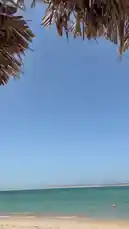 A serene beach scene under a clear blue sky, framed by the edge of a thatched umbrella. Gentle waves lap at the shore, creating a tranquil atmosphere. Overlaid with calming Arabic prayer music, the video evokes a sense of peace and reflection. by @ru.ab71 with يارب رحمتك وسعت كل شي 🍃🤎 by Na🍃🤎