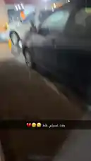 A nighttime scene during a heavy rainstorm, showing parked vehicles including a white pickup truck and a black car in a wet parking lot. Streetlights illuminate the area, reflecting off the rain-soaked ground. A building with Arabic signage is visible in the background. The camera pans across the scene, capturing the intensity of the downpour. by @by.sul with by.sul by Original Sound
