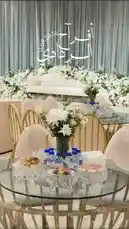 A tour of a beautifully decorated wedding reception hall, showcasing a grand stage with white floral arrangements, elegant seating areas with gold-trimmed tables, and a stunning dessert display. The video features a server presenting an array of elaborate desserts, including Ferrero Rocher towers, chocolate truffle pyramids, and colorful macarons, all set to upbeat Arabic music. by @me_ha73 with Original Sound by me_ha73