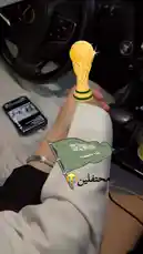 A close-up shot from inside a car shows a person's hands, one with red nail polish, holding a phone displaying a soccer game. Augmented reality filters overlay a golden FIFA World Cup trophy and a waving Saudi Arabian flag onto the scene, creating a festive atmosphere. The background features a car dashboard and steering wheel. A male voice sings a traditional Arabic song, adding to the celebratory mood. by @ab.7955 with Original Sound by ab.7955