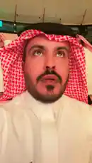 A man identified as Yaser Al-Khamsan, wearing traditional Saudi attire including a white thobe and red-and-white checkered ghutra, films himself and his guests at what appears to be a formal gathering or cultural event. He greets several men in similar traditional clothing, creating a warm and welcoming atmosphere. The setting is an indoor venue with tiered seating, where other attendees, including women in black abayas, are seated in the background. The camera pans across the room, capturing the social interaction before settling on a stage area with Arabic text on a large screen, suggesting an organized presentation or ceremony. by @y_alkhmsan with Original Sound by y_alkhmsan