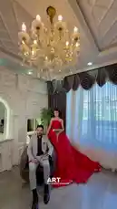 A beautifully styled photoshoot featuring a couple in formal attire within an opulent room. The woman wears a stunning red strapless gown with sparkling details and holds a bouquet of white flowers, while the man is dressed in a light gray suit. They pose together under a grand crystal chandelier, surrounded by elegant architectural details and luxurious curtains. This high-quality cinematic portrait captures a moment of romance and sophistication. by @artvideo.me with artvideo.me by Contains: Pitne Digel Te - Karwan Kamil