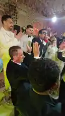A lively wedding reception is captured in this energetic video. Guests, including the bride and groom, are seen dancing enthusiastically on the dance floor to upbeat music. A man in a suit sings into a microphone, leading the celebration. The atmosphere is festive, with people cheering, clapping, and raising their hands in the air. by @mo.fouadziko with Original Sound by mo.fouadziko