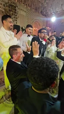 A lively wedding reception is captured in this energetic video. Guests, including the bride and groom, are seen dancing enthusiastically on the dance floor to upbeat music. A man in a suit sings into a microphone, leading the celebration. The atmosphere is festive, with people cheering, clapping, and raising their hands in the air. by @mo.fouadziko with Original Sound by mo.fouadziko