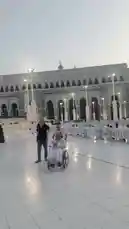 Experience the serene atmosphere of Masjid al-Haram in Mecca as a pilgrim in a wheelchair is gently pushed through the vast, white-tiled courtyard. The camera pans across the iconic architecture, capturing the grandeur of the mosque's minarets and the surrounding modern cityscape, including the towering Abraj Al-Bait complex. This peaceful moment highlights accessibility within one of Islam's holiest sites, while a prayer is recited in Arabic, adding a profound spiritual layer to the visual journey. by @om-5p with " الصلاة على النبي ﷺ . by -  💙📚🌿 " .