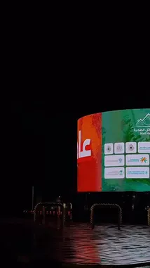 Experience a cinematic night drive through Riyadh, Saudi Arabia. The journey begins with a close-up of a massive, curved digital billboard displaying 'نجمة' and 'صحة' in Arabic, promoting Najd Health City. As the camera moves, it reveals a wider view of the wet, reflective road, illuminated streetlights, and a prominent red-and-white chevron sign guiding drivers along the curve. This atmospheric clip captures the modern urban landscape of Riyadh after dark. by @karim.alsaleh with مالك ديار تشابه قلب حايل ♥️ by 📍Hai’l