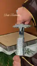 Watch as a person dressed in traditional Middle Eastern attire uses an ornate sword to cut a rectangular 'Date Tiramisu' cake. The dramatic reveal shows the layered dessert on a plate, topped with a miniature palm tree decoration. This unique and visually striking presentation combines culinary art with cultural flair. by @fox_cofee with Original Sound by fox_cofee