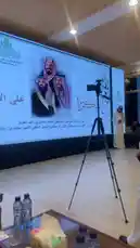 The video captures a formal event hosted by the Abdullah Rasool Al-Rasool Private Foundation For His Family in Riyadh. A man, identified as Yaser Al-Khamsan, delivers a speech at a podium in front of a large screen displaying Arabic text and images of Saudi royalty. The setting is a well-lit conference hall, and the speaker is dressed in traditional Saudi attire. by @y_alkhmsan with Original Sound by y_alkhmsan