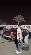 This nighttime scene captures a group of people working together to repair a dark-colored sedan on a paved area, possibly a racetrack or parking lot. The car has significant rear-end damage, and one person is actively changing a tire while others observe. The setting includes palm trees, concrete barriers, and distant city lights, creating a candid moment of roadside assistance under the night sky. by @mo_kbs8 with Original Sound by mo_kbs8